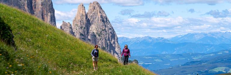 Two hikers walking along a green mountain trail in the Dolomites, part of the Sentiero del Castagno in South Tyrol