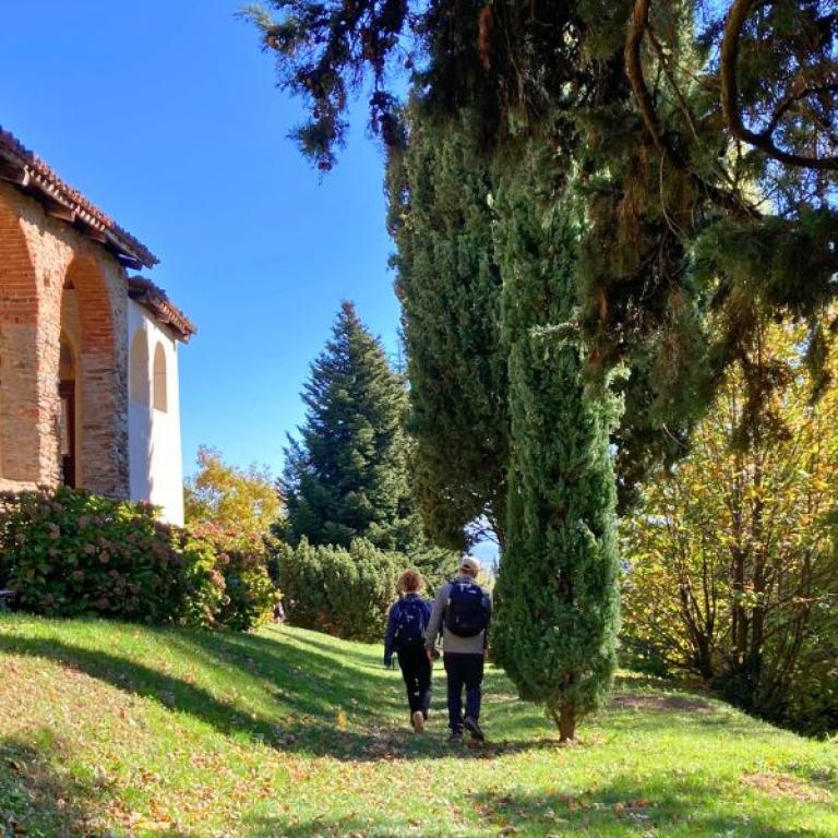 Due camminatori seguono il Cammino di Oropa accanto a una cappella di campagna immersa nel verde.